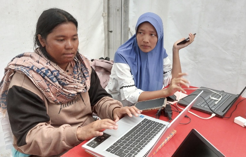 Eska Laurensia Sinabutar (berjaket coklat hitam), mengerjakan tugas akhir akademiknya di Pusat Informasi dan Media Center Kementerian Komunikasi dan Digital (Kemkomdigi) yang berlokasi di GOR Pandan, Tapanuli Tengah, Kamis (4/12/2025). (Foto: InfoPublik)